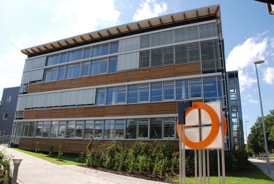 Moderner S-Bau der Hochschule Rosenheim mit großem Hochschullogo an der Fassade, Glasfront und Vorplatz bei Tageslicht