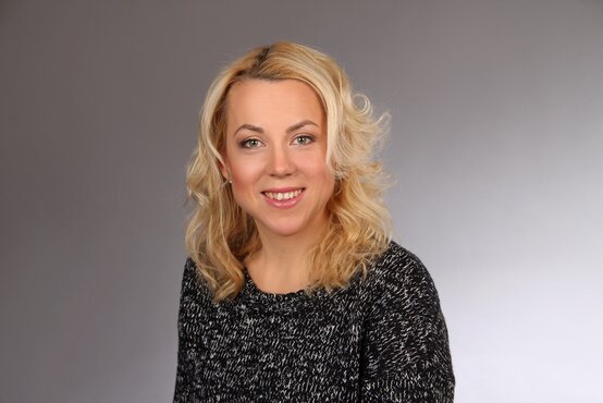 Yvonne Ortmeier. Porträt von lächelnde Frau mit lockigen blonden Haaren vor grauem Hintergrund