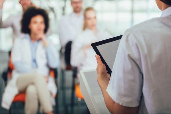 Person im weißen Kittel hält ein Tablet und spricht vor mehreren sitzenden Personen in hellem, modernem Raum