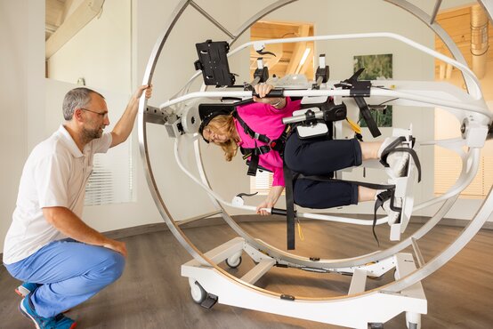 Person mit pinkfarbenem Oberteil und Gurten in ringförmigem Therapiegerät, zweite Person hält Metallring im hellen Raum fest