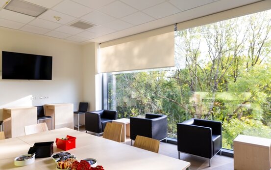 Heller Aufenthaltsraum im Krankenhaus mit Panoramafenstern, schwarzen Sesseln, Holztischen und Snacks auf dem Tisch