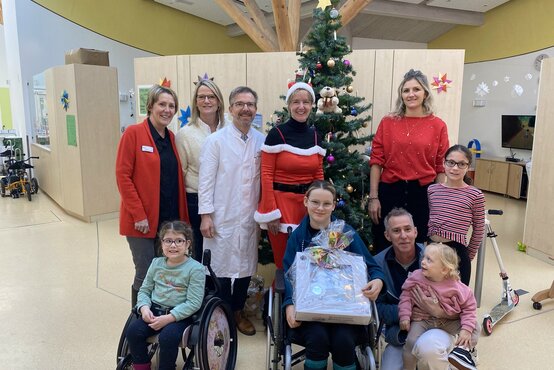 Gruppe aus Klinikpersonal und Kindern, teils im Rollstuhl, mit gespendetem Geschenk vor Weihnachtsbaum in Klinik
