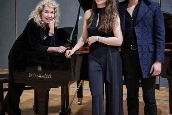 Drei Personen in eleganter Kleidung an schwarzem Konzertflügel im Musiksaal, Holzboden und Akustikwände im Hintergrund