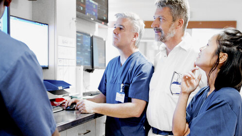 Vier Personen, drei in blauen Oberteilen, eine im weißen Hemd, stehen vor mehreren Monitoren an einer Arbeitsfläche