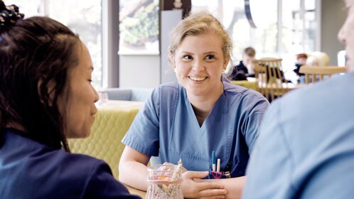 Drei Personen in blauer Arbeitskleidung sitzen an einem Holztisch in hellem Aufenthaltsraum mit weiteren Personen im Hintergrund