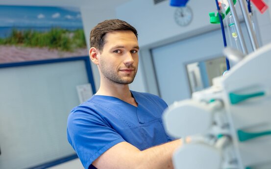 Junger Mann in blauer medizinischer Kleidung steht in hellem Klinikraum neben medizinischen Geräten