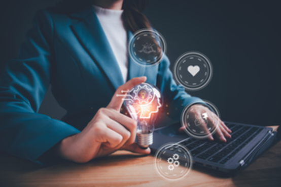 Person im blauen Blazer hält Glühbirne vor Laptop, umgeben von digitalen Symbolen auf Holztisch vor dunklem Hintergrund