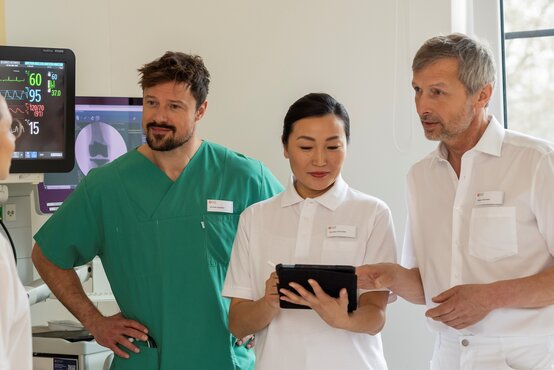 Vier medizinische Fachkräfte im Behandlungsraum, zwei in weißen Kitteln, eine in grüner OP-Kleidung, Tablet und Monitor im Hintergrund