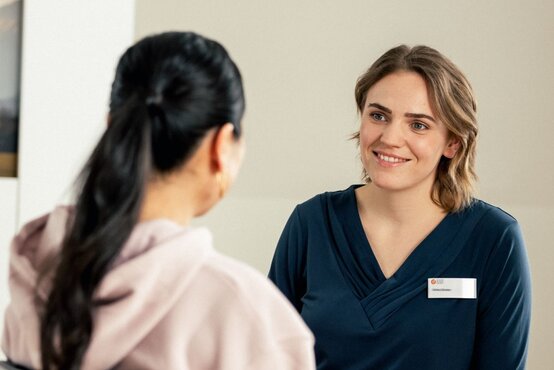 Zwei Frauen sitzen sich im freundlich eingerichteten Raum gegenüber, eine mit Namensschild in dunkelblauer Kleidung lächelt
