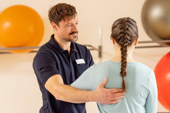 Physiotherapeut betreut Patientin mit geflochtenem Zopf im modernen Therapieraum, bunte Gymnastikbälle im Hintergrund