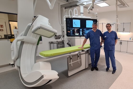 Zwei Ärzte in blauer OP-Kleidung stehen in einem modernen Eingriffsraum mit angiographischem Röntgengerät