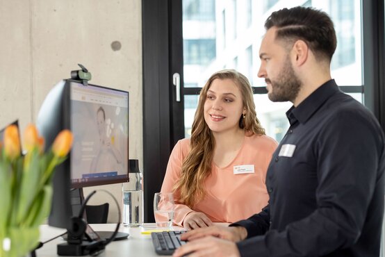 Frau in Bluse und Mann im dunkelblauen Hemd arbeiten gemeinsam am Computer im modernen Büro mit Tulpen und Glas Wasser