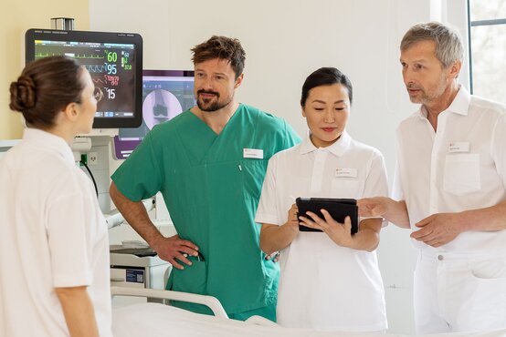 Vier Ärztinnen und Ärzte im Team besprechen sich auf der Krankenhausstation am Patientenbett mit Monitor und Tablet