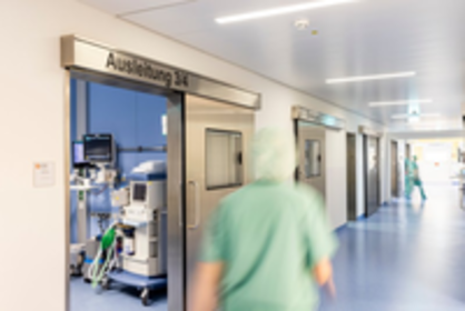 Krankenhausflur mit Schild „Anästhesie 1“, medizinische Geräte im Raum, zwei Personen in grüner OP-Kleidung im Flur