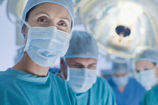 Mehrere Mediziner tragen OP-Kleidung, OP-Masken und Hauben, stehen unter OP-Lampen in einem Operationssaal und schauen in Richtung Kamera