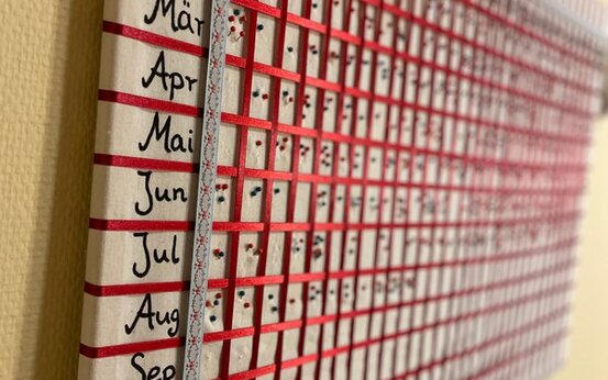 Selbstgebastelter Kalender an gelber Wand mit Monaten Januar bis Dezember, Tabelle mit Zahlen 1 bis 31, rote Linien als Raster mit Markierungen