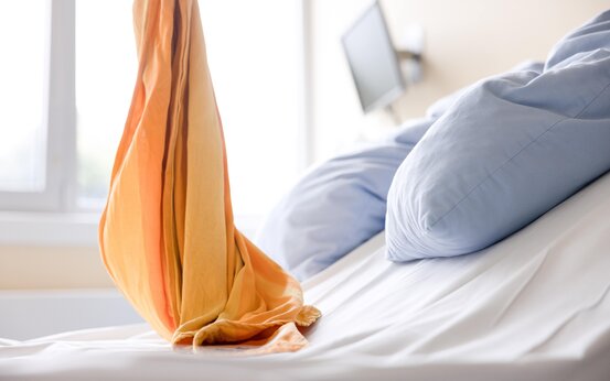 Heller Kreißsaal mit Bett, weißem Bettlaken, hellblauem Kissen und orange-gelber Decke, Fenster und Wandmonitor im Hintergrund