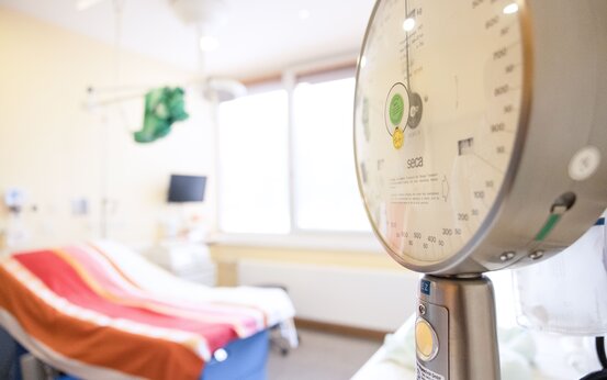 Rundes medizinisches Messgerät im Vordergrund, Krankenhausbett mit bunter Decke und Monitor im Hintergrund