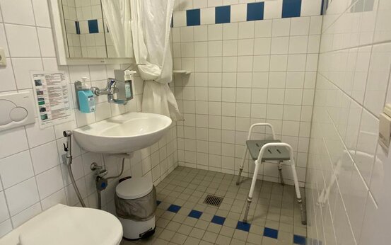 Barrierefreies Badezimmer mit weißen Fliesen, blauen Akzenten, Spiegel, Waschbecken, wandhängendem WC und Duschstuhl 