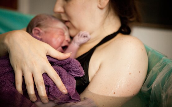 Erwachsene Person hält Neugeborenes auf Oberkörper, Baby in lila Handtuch, beide im Wasserbecken 