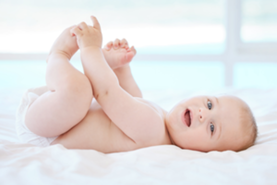 Baby liegt auf dem Rücken auf weißem Bett, hält die Füße mit den Händen, trägt eine Windel, unscharfer Hintergrund