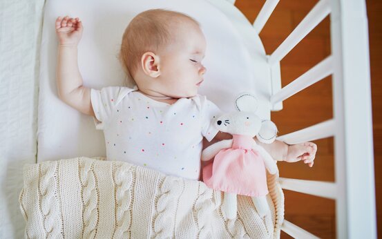 Schlafendes Baby im weißen Gitterbett, weißer Body mit bunten Punkten, hält Stoffmaus im rosa Kleid