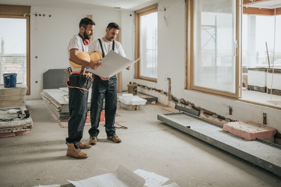 Zwei Bauarbeiter mit Bauplan und Schutzhelmen besprechen Arbeiten im hellen Rohbau einer Baustelle