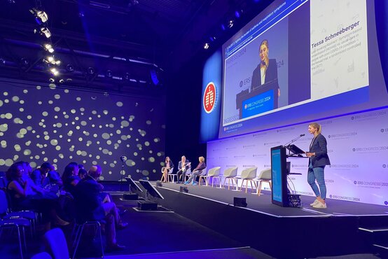 Referentin Tessa Schneeberger hält Vortrag auf moderner Bühne mit Publikum und Podium