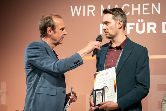 Zwei Männer vor brauner Wand mit Schriftzug, einer hält Mikrofon, der andere Urkunde