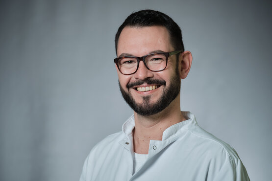 Portrait eines lächelnden Arztes mit dunklem Haar, Vollbart und Brille im weißen Arztkittel vor grauem Hintergrund