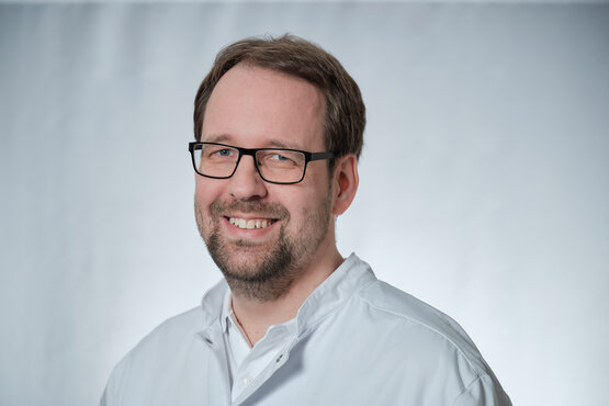 Portrait eines Arztes mit Brille im weißen Arztkittel vor grauem Hintergrund