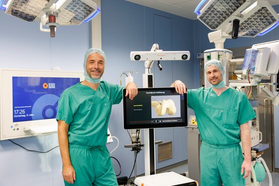 Zwei Chirurgen in grüner OP-Kleidung vor 3D-Monitor mit Knochen im modernen Operationssaal