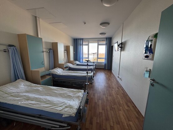 Krankenhaus-Mehrbettzimmer mit vier Betten, Vorhängen, Nachttischen, großem Fenster, Fernseher, Desinfektionsmittelspender an der Tür