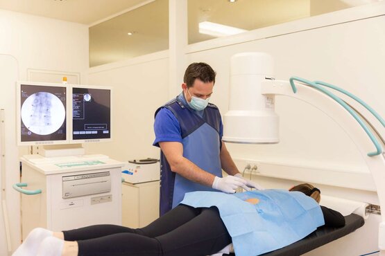 Arzt mit Mundschutz in blauer Schutzkleidung tastet den Rücken einer Patientin vor interventioneller Wirbelsäulenuntersuchung ab