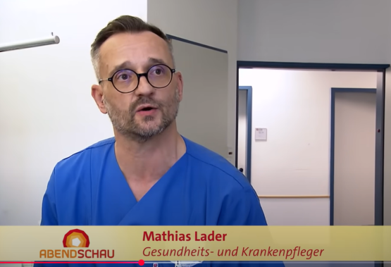 Mann mit kurzem Haar in blauer medizinischer Kleidung, gelbes Banner „ABENDSCHAU Mathias Lader Gesundheits- und Krankenpfleger“