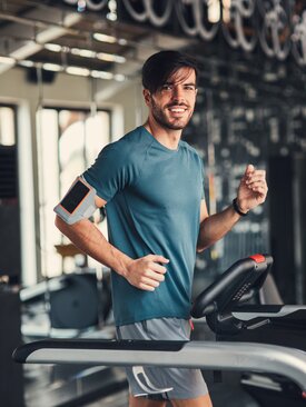 Junger Mann in blauem T-Shirt joggt lächelnd auf Laufband im Fitnessstudio