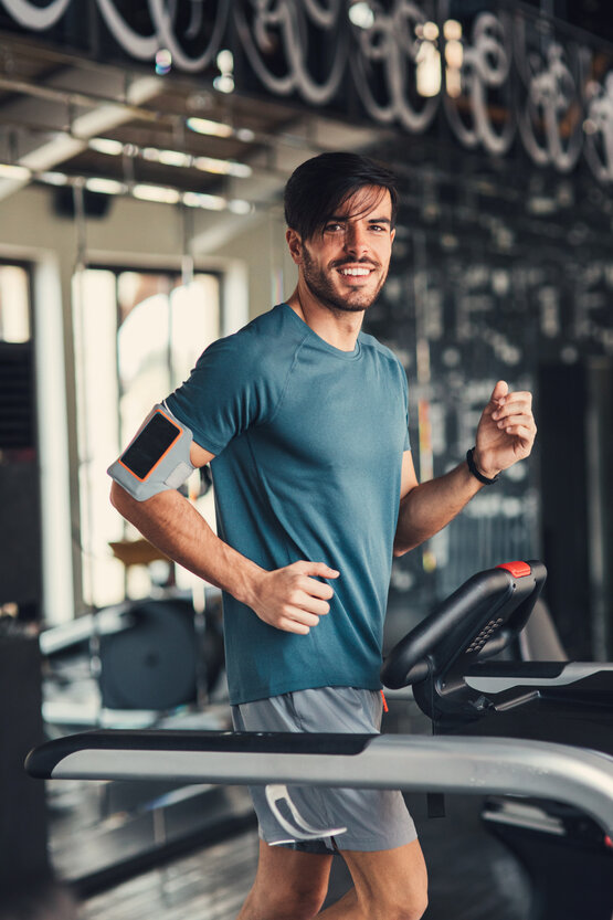Junger Mann in blauem T-Shirt joggt lächelnd auf Laufband im Fitnessstudio