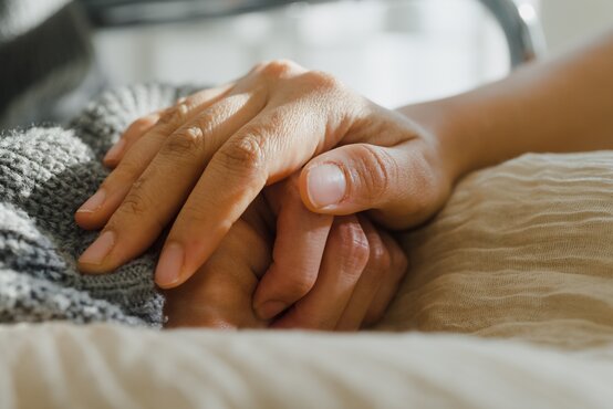 Nahaufnahme von Händen, die sich fürsorglich halten, mit grauer Strickdecke im Hintergrund.