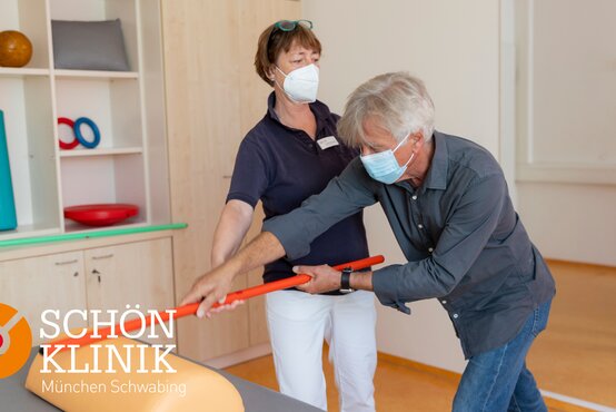 Zwei Personen mit Mundschutz üben im Therapieraum mit Gymnastikstab, Schön Kliniik München Schwabing Logo unten links