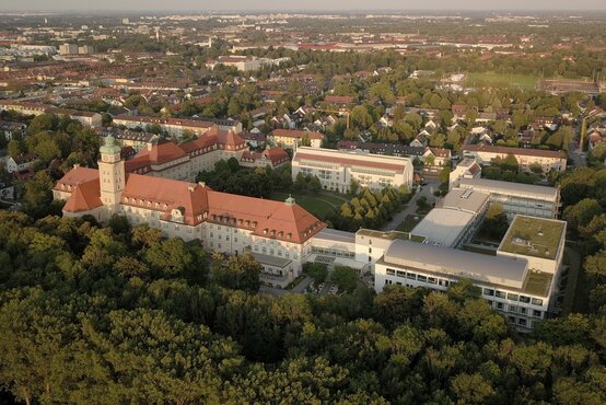Luftaufnahme der Schön Klinik Harlaching mit roten Dächern und Turm, umgeben von Parklandschaft und Wohnhäusern 